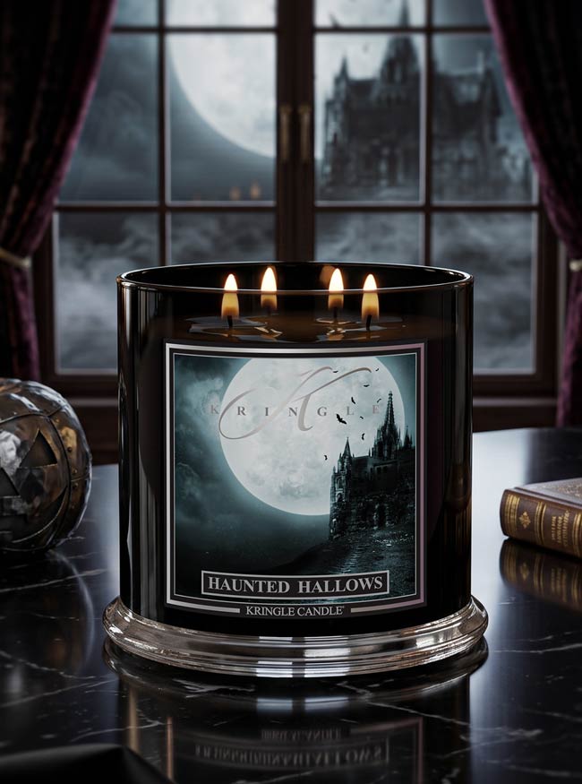 A black three-wick candle labeled 'Haunted Hallows' by Kringle Candle, set on a dark reflective table with a gothic window, moon, and castle visible in the background.