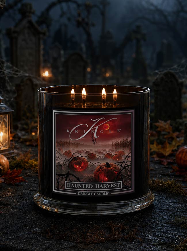 A  4-wick lit black glass jar candle with labeled 'Haunted Harvest' by Kringle Candle, set in a spooky graveyard scene with tombstones, lanterns, and autumn leaves.