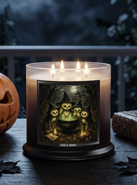 A lit three-wick candle in a dark amber glass jar sits on a wooden table outdoors at night. The front label shows a spooky illustration of three owl-like figures around a cauldron under a full moon, with the text 'OWL'S BREW' below.