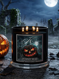 A black Kringle Candle jar labeled 'Pumpkin Head' with four lit wicks, shown on a stone surface in a spooky Halloween setting with a carved pumpkin and full moon in the background.