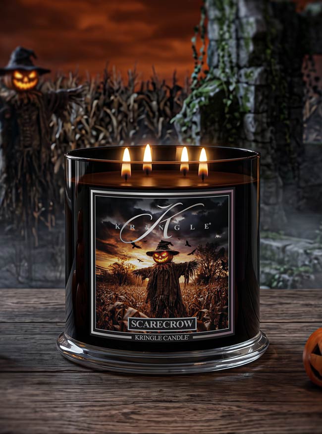 A black glass jar candle with four lit wicks sits on a wooden surface. The front label shows a scarecrow in a field under a dark sky, with the words 'Kingle' and 'SCARECROW' visible.