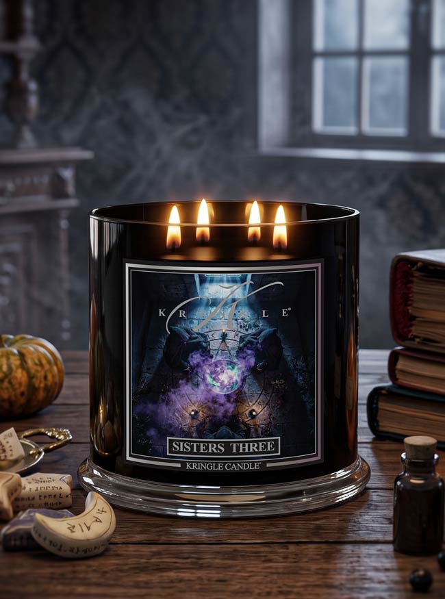 A black glass jar candle with three lit wicks and glowing flames, labeled 'Kringle Candle Sisters Three,' set on a rustic wooden table with occult-themed props like a small pumpkin, books, and a bottle in a dimly lit room.