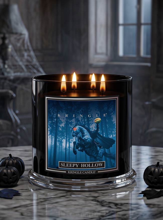 A black glass jar candle with four lit flames, featuring a blue forest label with a witch riding a broomstick and the text 'Kringle Sleepy Hollow.' The candle sits on a marble surface with small jack-o'-lantern props, against a dark moody interior background.