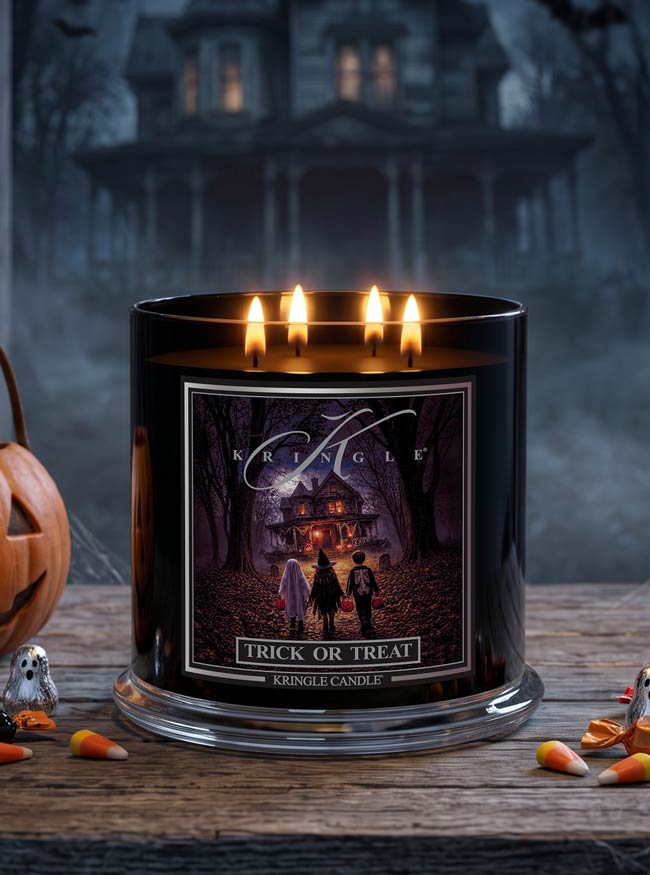 A black glass jar candle with four lit flames sits on a rustic wooden surface. The front label shows a haunted-house illustration with the text 'Kringle' and 'Trick or Treat'.