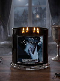 A black glass jar candle with four lit wicks and a gothic label reading 'K Agle' and 'Vampire's Kiss', set on a wooden table with a blurred window and silver goblet in the background.