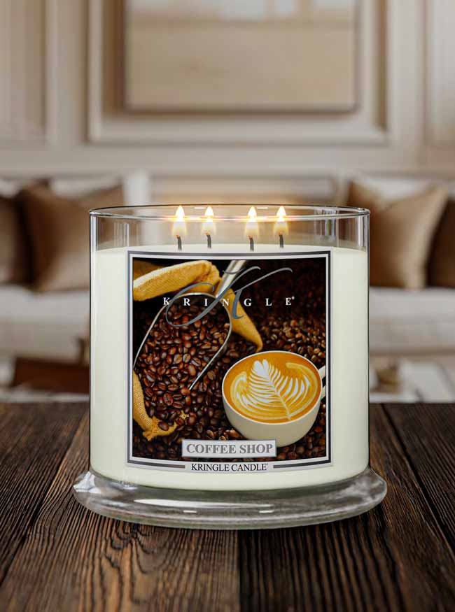 A lit Kringle Candle in a clear glass jar sits on a dark wooden table in a softly blurred living room setting. The candle label features coffee-themed imagery, including coffee beans, a cup of latte, and a biscuit, with the text 'KRINGLE CANDLE' and 'COFFEE SHOP'.