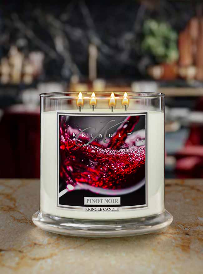 A four-wick Kringle Candle in a clear glass jar with white wax and lit flames, featuring a front label with a red wine glass graphic and the scent name 'Pinot Noir'.