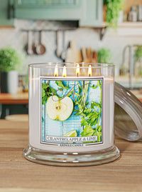 A lit Kringle Candle in a clear glass jar with a glass lid resting beside it on a wooden table. The candle features a white wax body and a colorful front label depicting a sliced apple, lime, and green herbs on a blue background, with the scent name 'Cilantro, Apple & Lime' and the brand 'KRINGLE CANDLE' printed below.