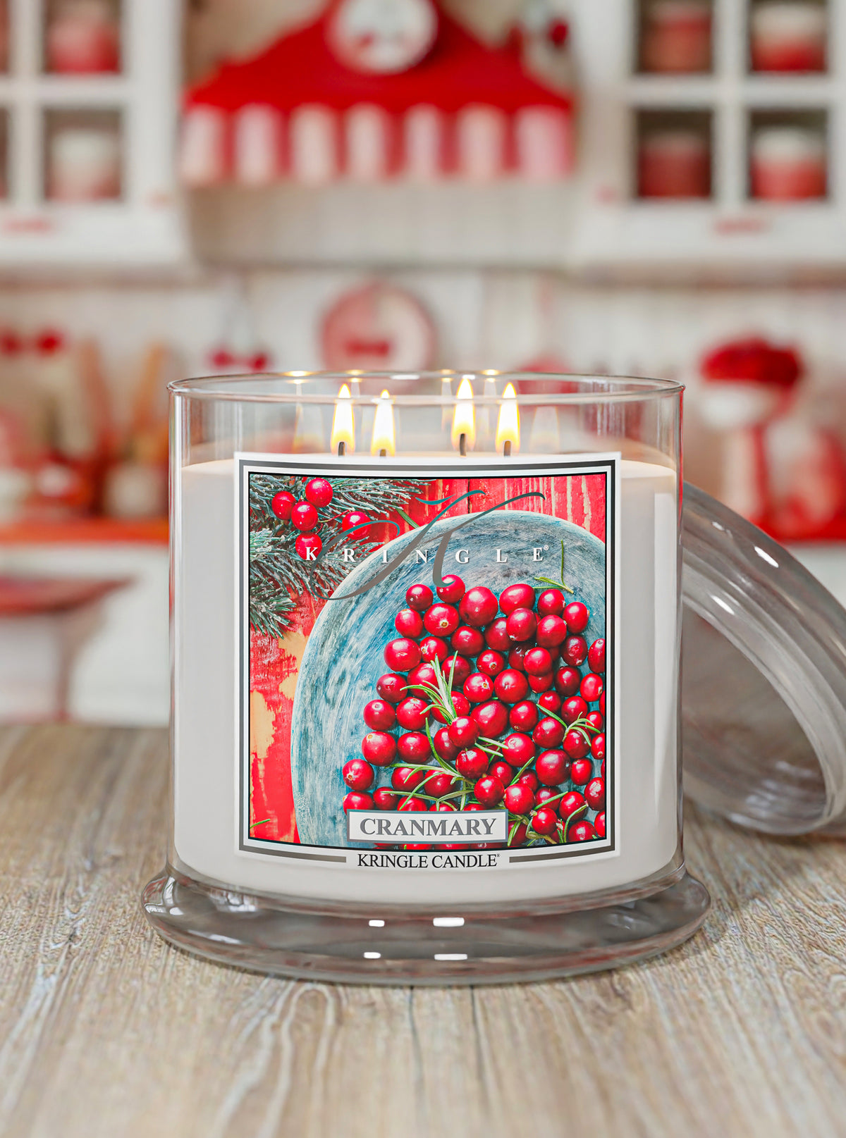 A lit white scented candle in a clear glass jar with a lid resting beside it on a wooden surface. The front label shows a red cranberry illustration and the text 'CRANMARY' and 'KINGLE CANDLE.