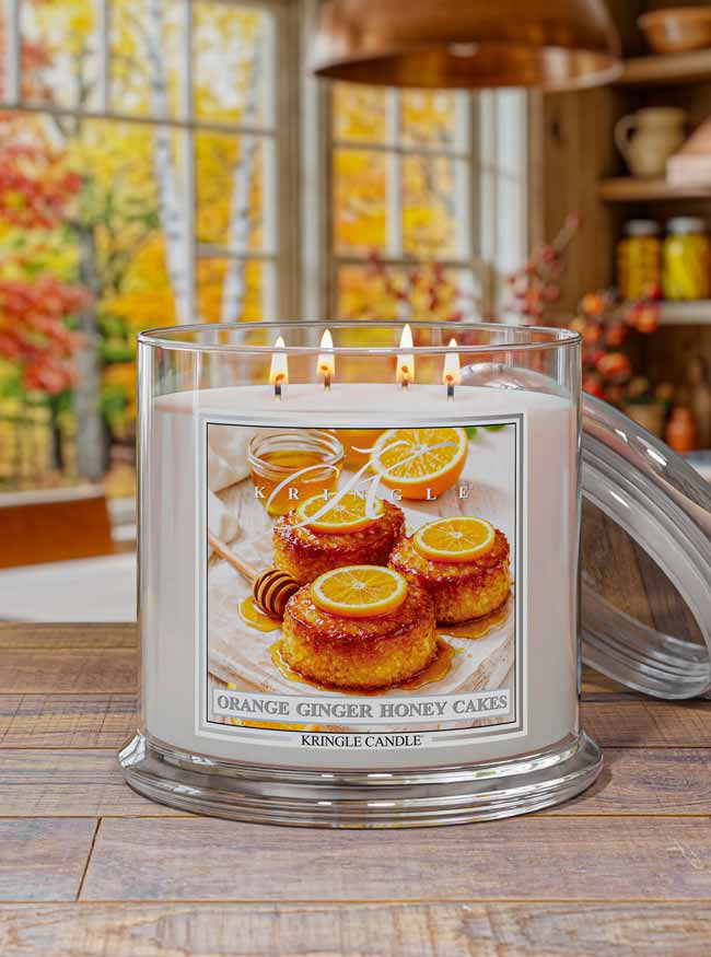 Kringle Candle with 'Orange Ginger Honey Cakes' label on a wooden surface with autumn decor in the background.
