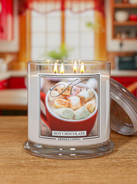 A lit Kingle Candle jar labeled 'Hot Chocolate' in a clear glass container with a lid resting beside it on a wooden table. The candle wax is white and the label shows a mug of hot chocolate with marshmallows.