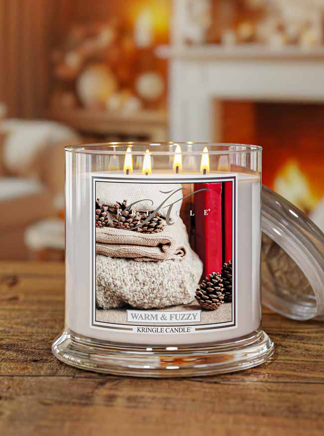 A lit Kringle Candle in a clear glass jar sits on a wooden table with its glass lid leaning against the right side. The candle has white wax and a front label showing a cozy image of folded beige knit fabric, pinecones, and a red book, with the scent name 'Warm & Fuzzy'.