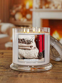 A lit Kringle Candle in a clear glass jar sits on a wooden table with its glass lid leaning against the right side. The candle has white wax and a front label showing a cozy image of folded beige knit fabric, pinecones, and a red book, with the scent name 'Warm & Fuzzy'.