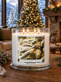 A large glass jar candle with four lit wicks sits on a wooden table in a cozy holiday living room. The candle label reads 'Black Forest Balsam' and shows snowy pine branches.