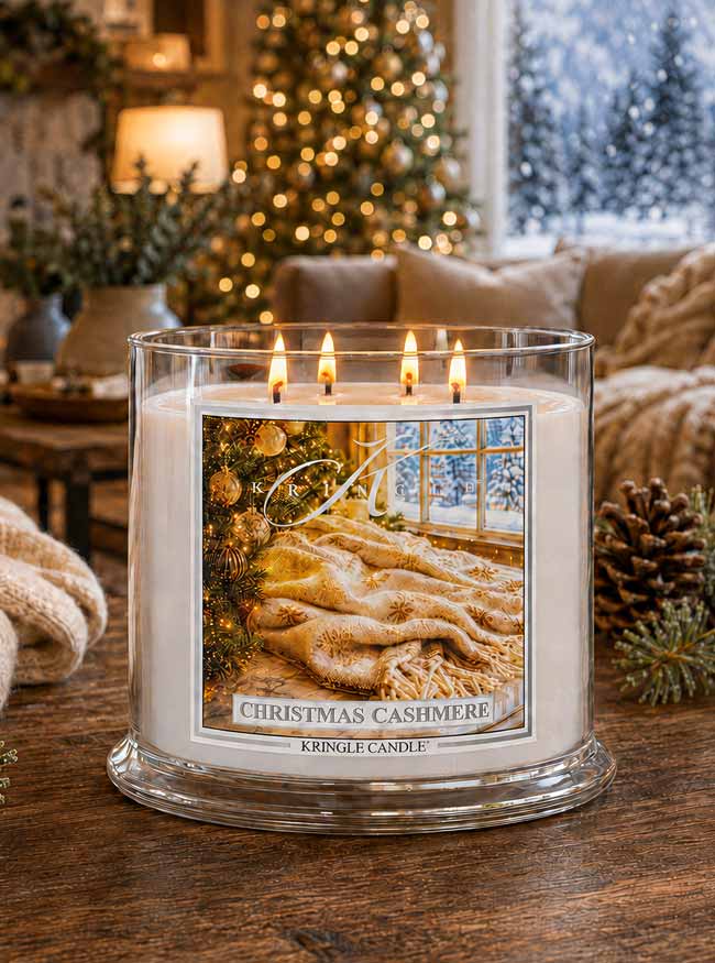 A lit Kingle Candle in a clear glass jar sits on a wooden table in a cozy living room setting. The candle features a square label with a warm-toned illustration of a cozy bed and window, and the text 'CHRISTMAS CASHMERE' and 'KINGLE CANDLE'.