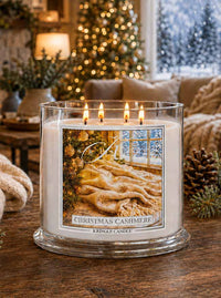 A lit Kingle Candle in a clear glass jar sits on a wooden table in a cozy living room setting. The candle features a square label with a warm-toned illustration of a cozy bed and window, and the text 'CHRISTMAS CASHMERE' and 'KINGLE CANDLE'.