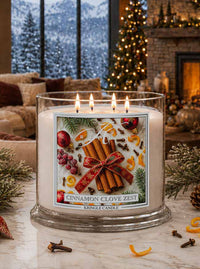A large three-wick scented candle in a clear glass jar sits on a marble surface in a cozy living room. The candle label reads 'CINNAMON CLOVE ZEST' and 'KRINGLE CANDLE,' with festive illustrations of cinnamon sticks, cloves, berries, citrus slices, and evergreen branches.