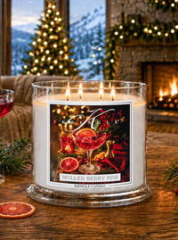 A large glass jar candle with four lit wicks sits on a wooden table in a cozy holiday living room. The candle label reads 'KRINGLE CANDLE' and 'MULLED BERRY PINE,' with festive artwork showing a red mulled wine glass, berries, citrus slices, and holiday decorations.