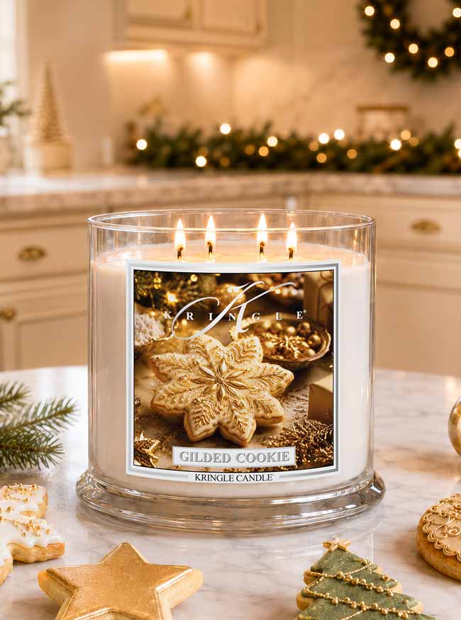 A lit Kingle Candle in a clear glass jar sits on a marble countertop in a warm, decorated kitchen. The candle label reads 'Gilded Cookie' and features an image of a gilded cookie.