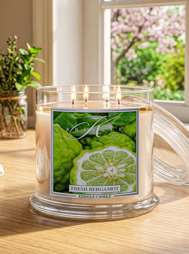 A Kringle Candle in a clear glass jar with three lit wicks, labeled 'Fresh Bergamot,' sitting on a wooden table with a window and plants in the background.