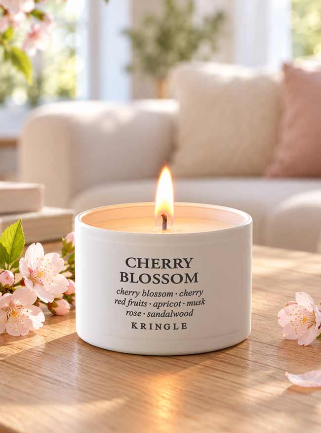 A lit white candle tin labeled 'CHERRY BLOSSOM' by Kringle sits on a wooden table in a softly blurred living room setting, surrounded by pink blossoms.