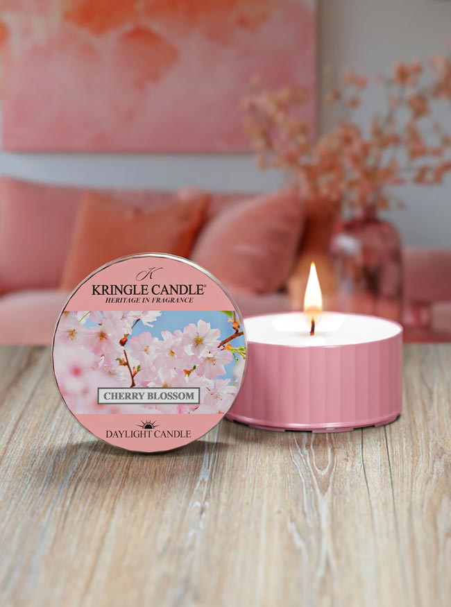 Pink candle  container with cherry blossom design on packaging, burning on a wooden surface.