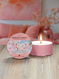 Pink candle  container with cherry blossom design on packaging, burning on a wooden surface.