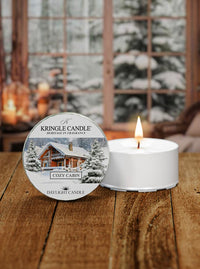 Kringle Candle Cozy Cabin candle on a wooden surface with a snowy cabin background
