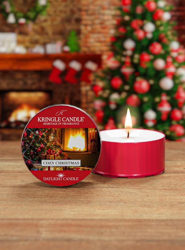 Kringle Candle Cozy Christmas daylight candle with packaging on a wooden surface, Christmas tree and fireplace in the background