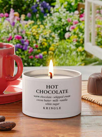 A lit white candle tin labeled 'HOT CHOCOLATE' by Kringle sits on a wooden table with a red mug on the left and a chocolate bar on the right, set against a blurred garden background.