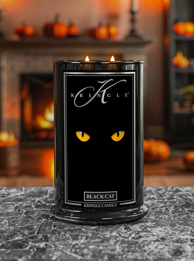 Kringle Candle Black Cat candle on a surface with pumpkins and a fireplace in the background