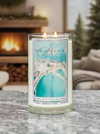Kringle Candle in a decorative glass jar on a marble surface with a fireplace and Christmas tree in the background.