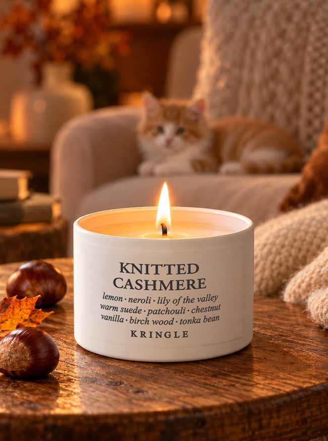 A lit white candle tin labeled 'KNITTED CASHMERE' by KRINGLE sits on a wooden table in a cozy indoor setting, with an orange cat resting on a sofa in the warm background.