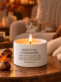 A lit white candle tin labeled 'KNITTED CASHMERE' by KRINGLE sits on a wooden table in a cozy indoor setting, with an orange cat resting on a sofa in the warm background.