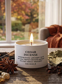 A lit white candle tin labeled 'CEDAR INCENSE' by KRINGLE sits on a rustic wooden table with star anise, cloves, and dried leaves, in front of a softly blurred window and cozy chair.