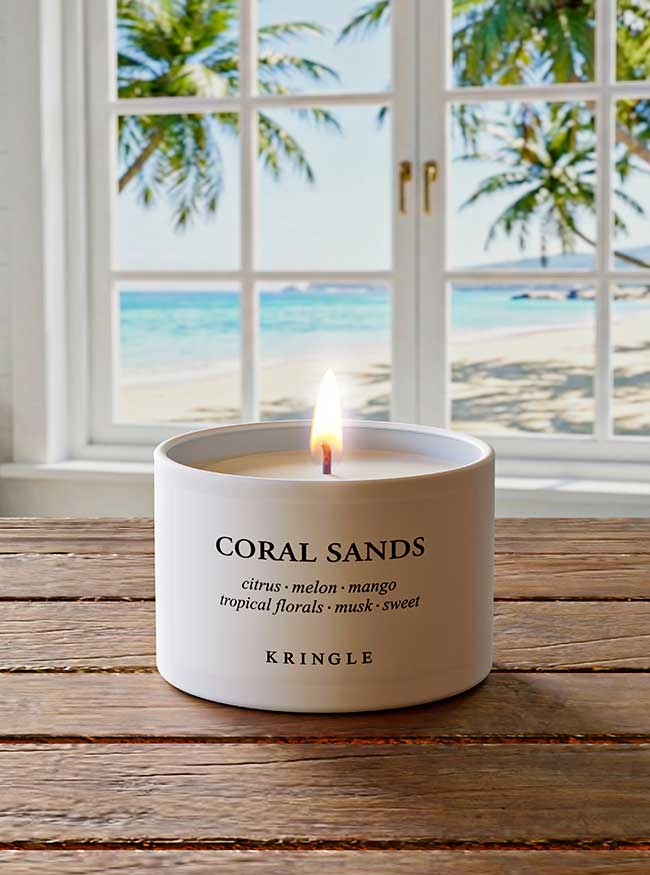 Kringle Candle labeled 'Coral Sands' on a wooden surface with a beach view through a window.