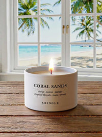 Kringle Candle labeled 'Coral Sands' on a wooden surface with a beach view through a window.