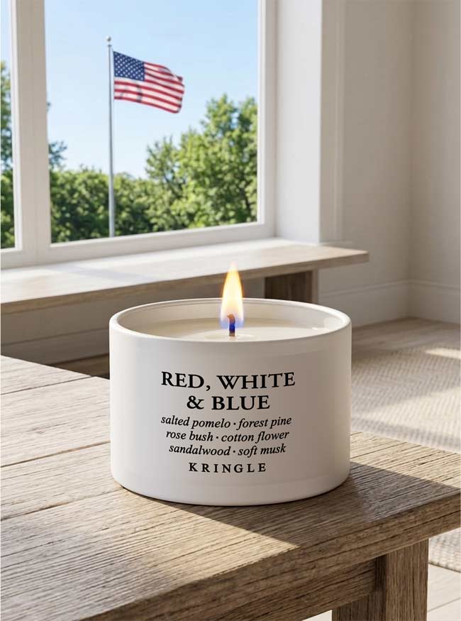 White candle labeled 'Red, White & Blue' on a wooden table with a window and American flag in the background.