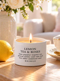 A lit white scented candle tin labeled 'LEMON TEA & ROSES' by KRINGLE sits on a wooden table in a softly styled interior setting with a lemon, flowers, and a teacup visible.