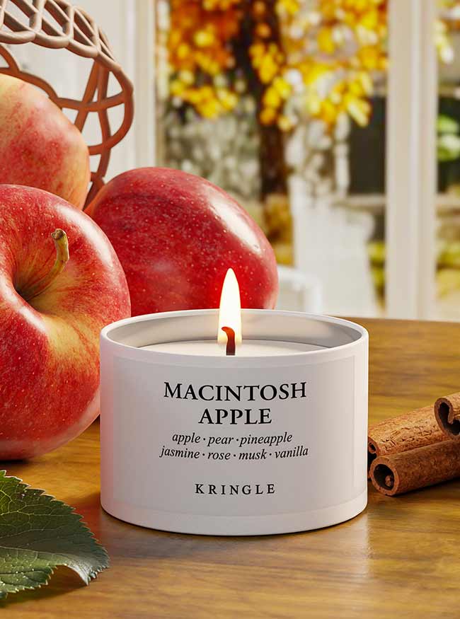 A lit Kringle Macintosh Apple scented candle in a white cylindrical tin, styled on a wooden table with fresh red apples, a cinnamon stick, and a leaf, with a blurred window and autumn foliage in the background.