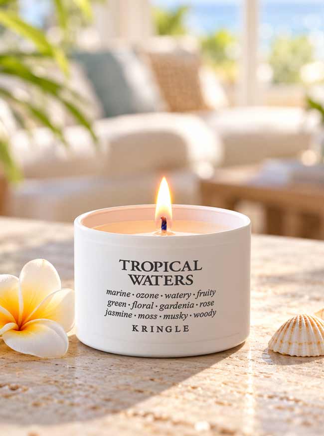 A lit white candle tin labeled 'TROPICAL WATERS' by Kringle sits on a sunlit stone surface with a plumeria flower and a small seashell nearby. The background is softly blurred, showing an outdoor patio or lounge setting.