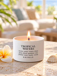 A lit white candle tin labeled 'TROPICAL WATERS' by Kringle sits on a sunlit stone surface with a plumeria flower and a small seashell nearby. The background is softly blurred, showing an outdoor patio or lounge setting.
