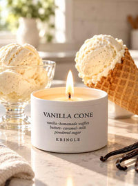 A lit white candle tin labeled 'VANILLA CONE' by Kringle sits on a marble surface, surrounded by vanilla ice cream scoops in a glass bowl and a waffle cone, with vanilla beans in the foreground.