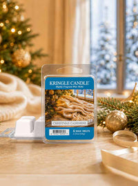 A package of Kringle Candle 'Christmas Cashmere' lightly fragrant wax melts sits on a tabletop with holiday decorations, including a blurred Christmas tree and ornaments, in the background.