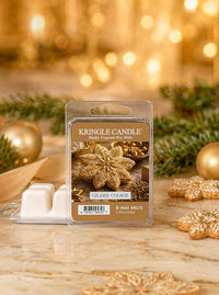 A package of Kringle Candle 'Gilded Cookie' scented wax melts stands on a marble surface with holiday decorations, including pine branches, gold ornaments, and cookie-shaped props, against a warm golden bokeh background.