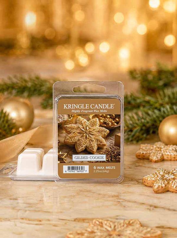 A package of Kringle Candle 'Gilded Cookie' scented wax melts stands on a marble surface with holiday decorations, including pine branches, gold ornaments, and cookie-shaped props, against a warm golden bokeh background.