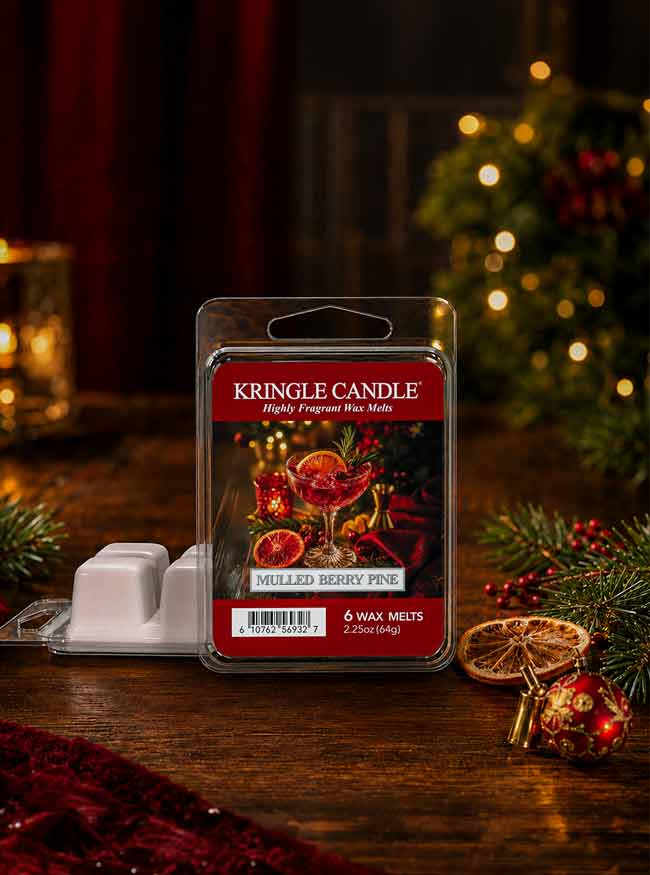 A package of Kringle Candle 'Milled Berry Puck' fragrance wax melts stands on a wooden table with holiday decorations in the background, including evergreen branches, red ornaments, and warm string lights.