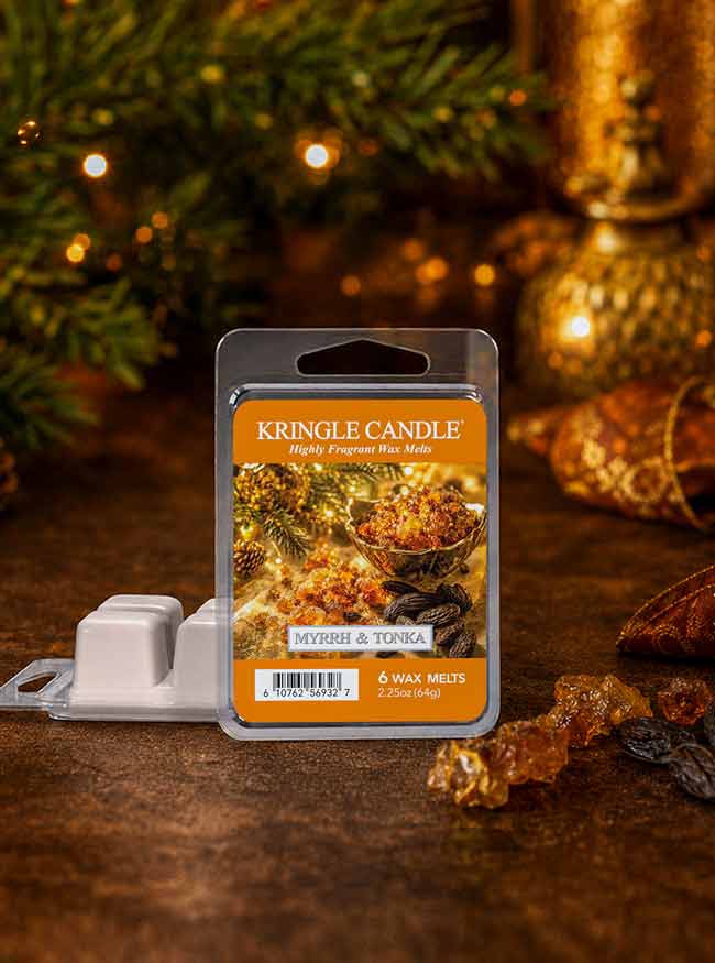 A package of Kringle Candle scented wax melts stands on a rustic wooden surface with holiday decor in the background. The clear plastic clamshell contains six wax melts labeled 'MYRRH & TONGA' and includes a small white plastic tray in front.