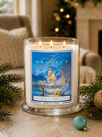 A lit Kringle Candle in a clear glass jar with a winter-themed label reading 'Bavarian Christmas.' The candle is styled on a tabletop with pine branches, ornaments, and a softly blurred indoor holiday background.