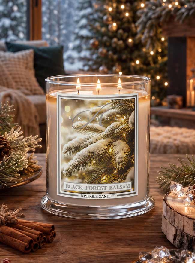 A lit Kringle Candle in a clear glass jar sits on a wooden table in a cozy holiday living room. The candle label features an illustration of snowy pine branches with warm lights, and the text reads 'BLACK FOREST BALSAM' and 'KRINGLE CANDLE'.